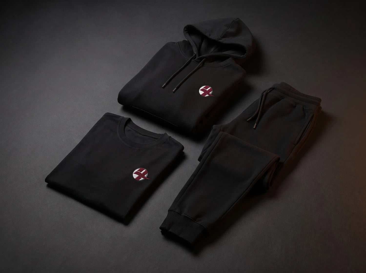 A black hoodie, sweatshirt, and jogger pants set arranged on a dark gray background. Each garment features a small circular red and white logo on the left chest.